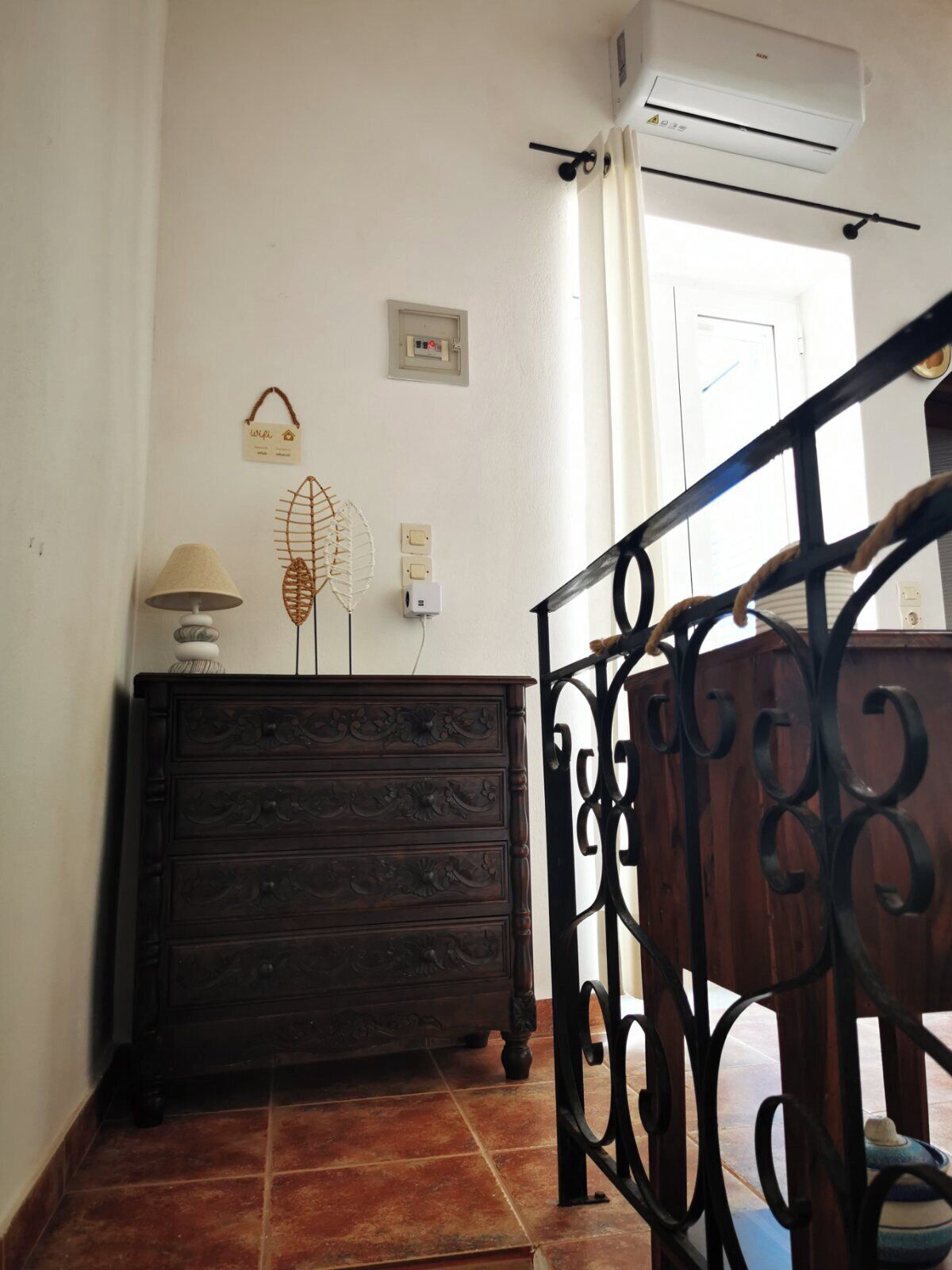 A room with a dark wooden dresser, a lamp, decorative wall hangings, an air conditioning unit, and a staircase with a metal railing.