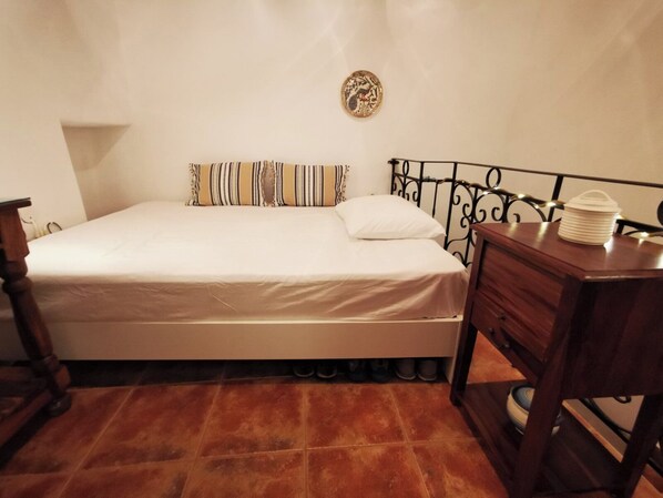 A bedroom with a bed, a wooden nightstand, a small table, and a wall-mounted decorative plate. - Authentic White House in Paros Lefkes (Paros)