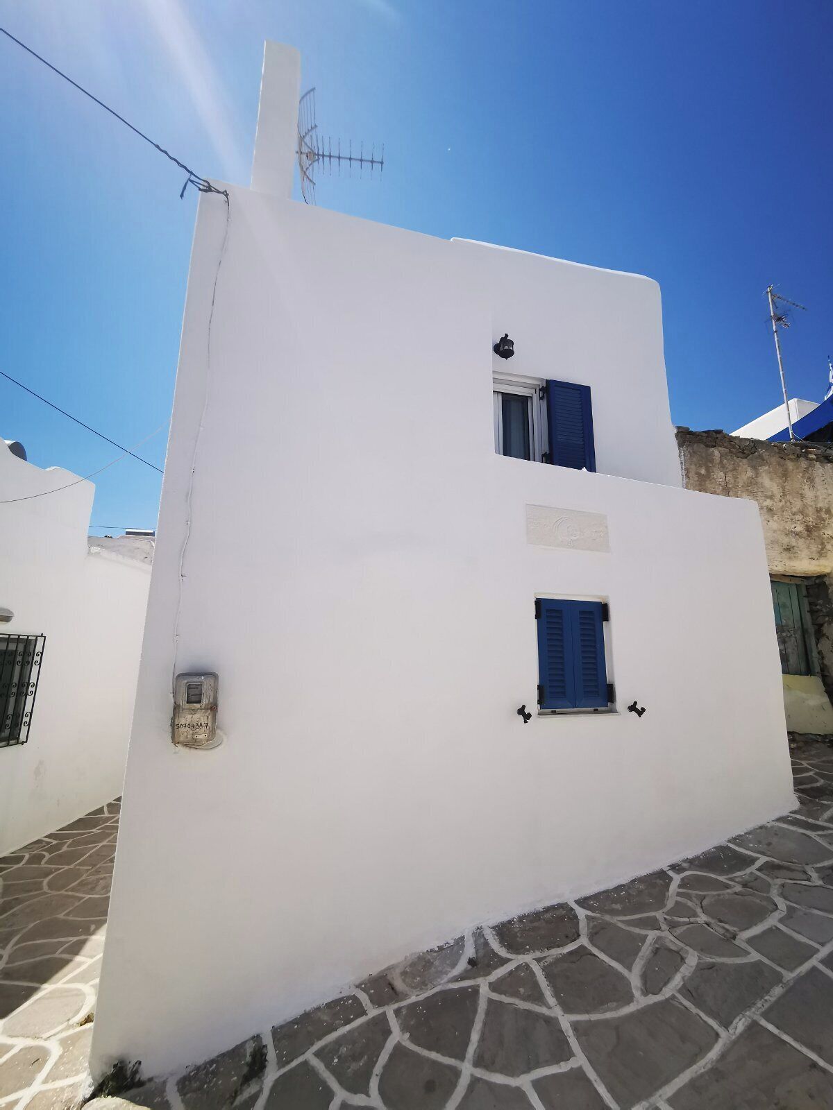 Um edifício branco com uma pequena janela, uma porta, antena parabólica e chão pavimentado em pedra.