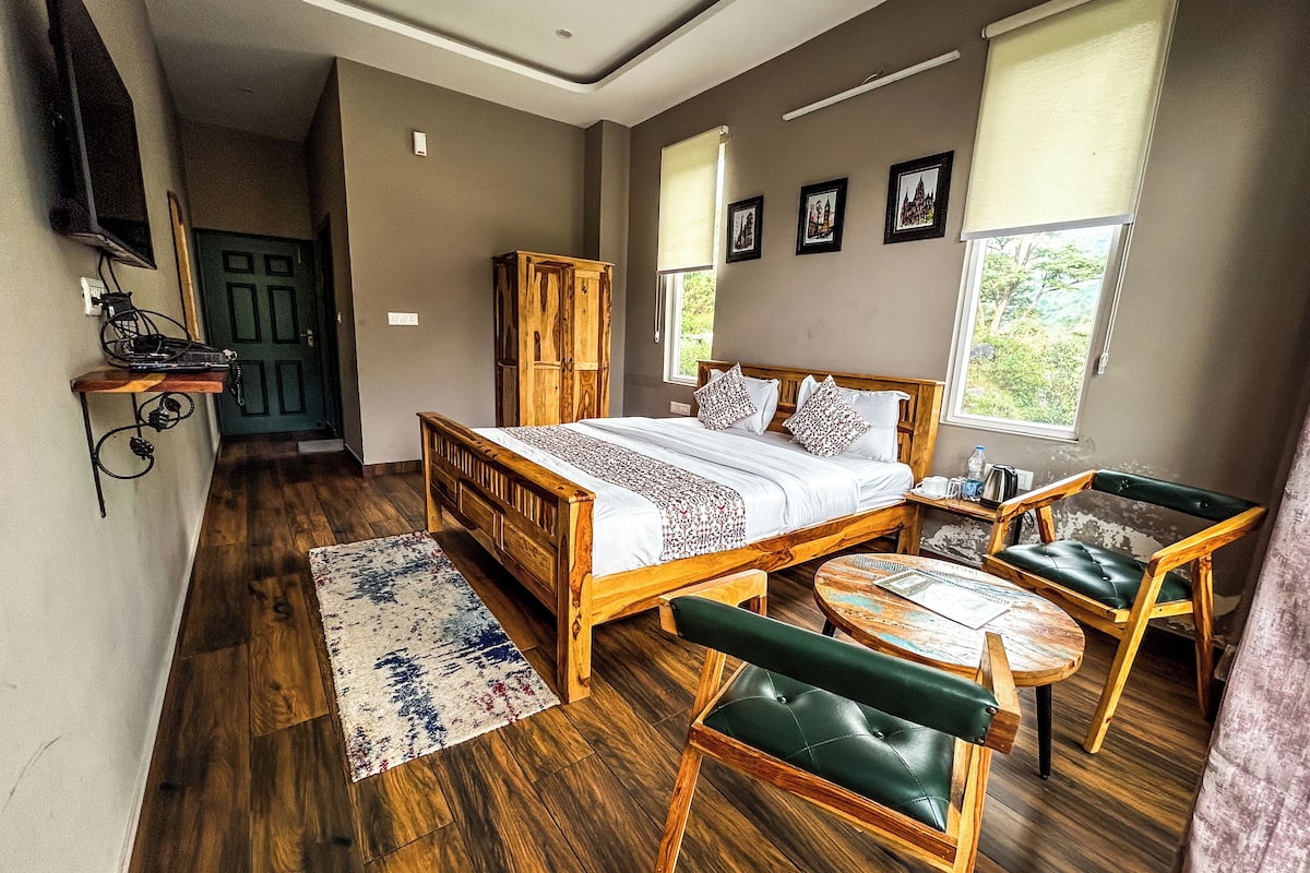 A bedroom with a wooden bed, a wooden chair, a small table, and a window with a view of greenery.