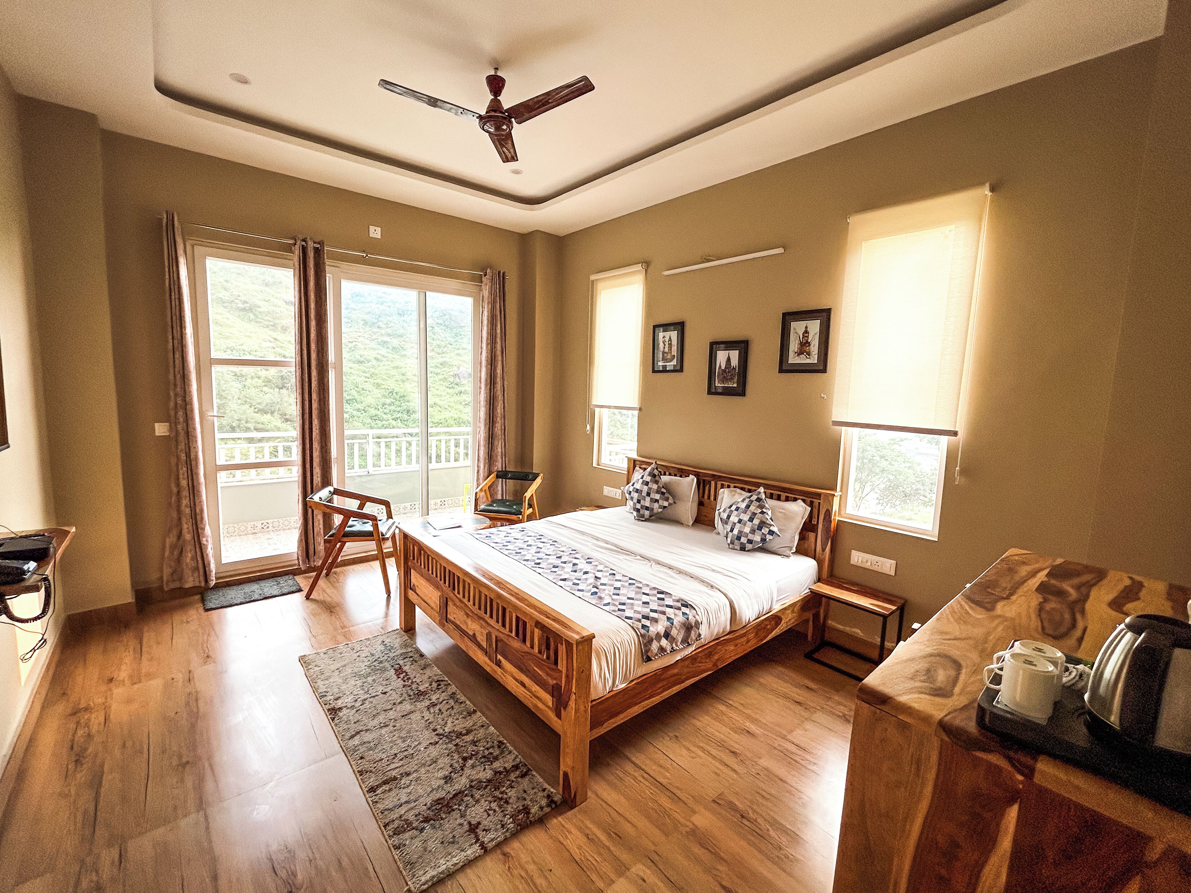 Une chambre spacieuse avec un grand lit, une tête de lit en bois et un ventilateur de plafond. Elle comprend un balcon avec des rideaux, un bureau avec une chaise et une machine à café sur la table de chevet.