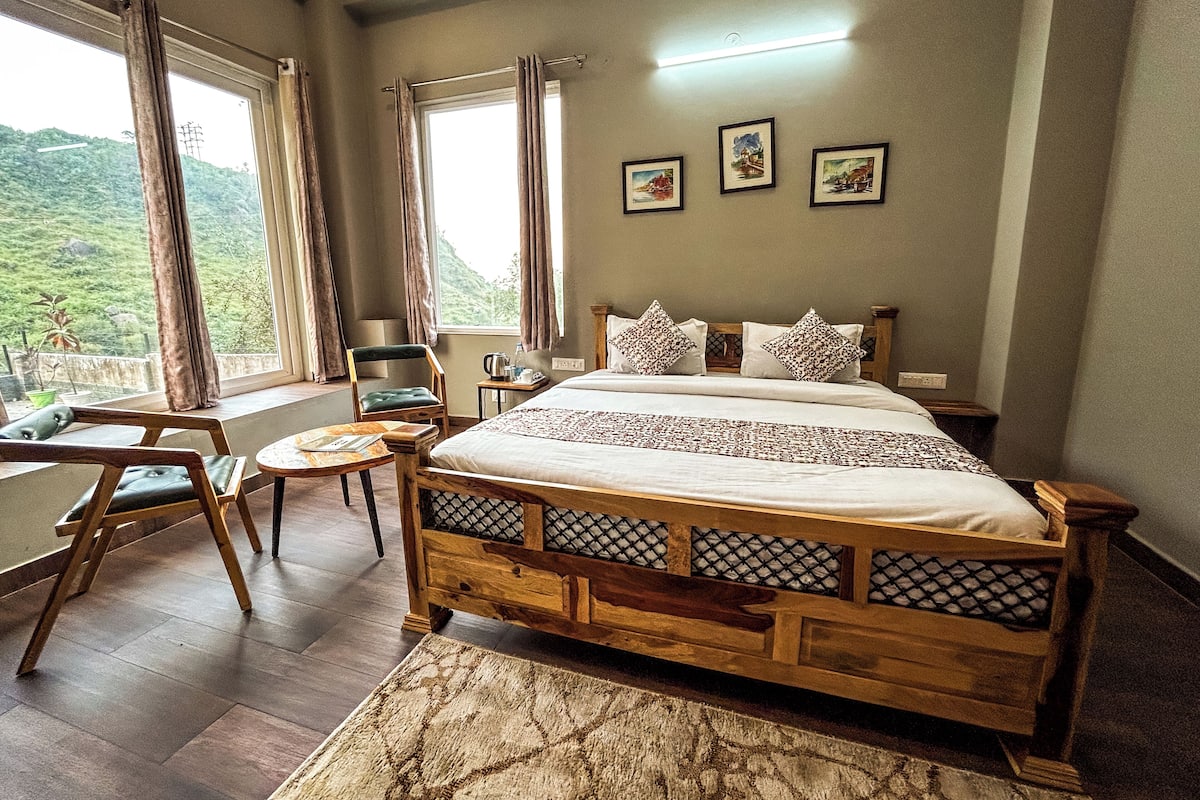 A bedroom with a large bed, a wooden headboard, a small table, two chairs, and a view of a mountainous landscape through the window.