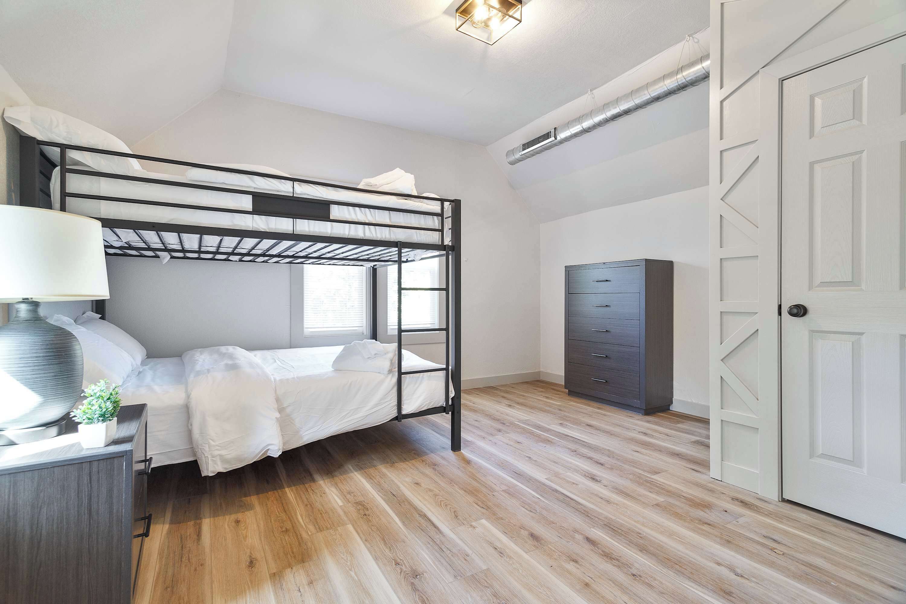 A bunk bed with a desk and lamp, a dresser, and a wardrobe in a room with wooden flooring and white walls.