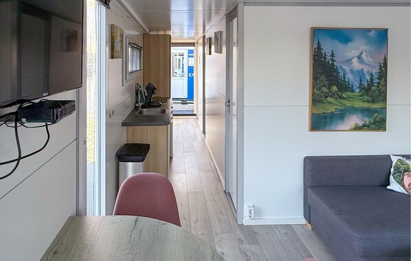 En smal korridor leder till ett litet kök med en diskho och en liten papperskorg. En tavla med ett landskapsmotiv hänger på väggen, och det finns en grå soffa i rummet.