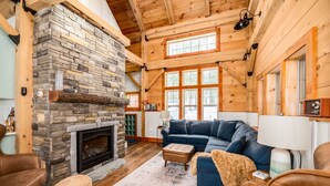 A cozy cabin interior with a stone fireplace, wooden beams, a blue sofa, and a large window.