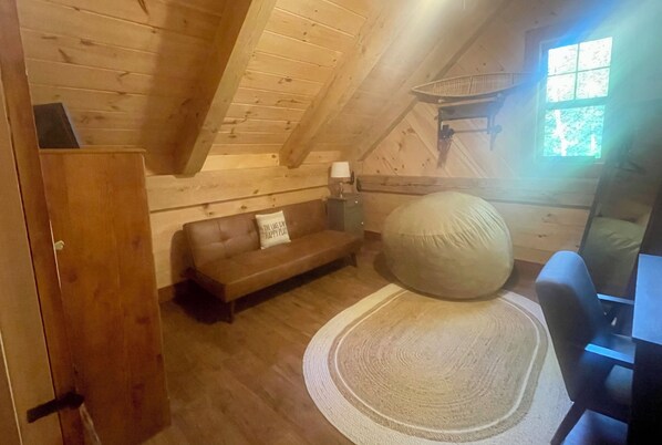 Une pièce chaleureuse avec des murs et un plafond en bois, un canapé marron avec un coussin, un fauteuil en forme de poire, une petite table avec une lampe et une fenêtre donnant sur des arbres.