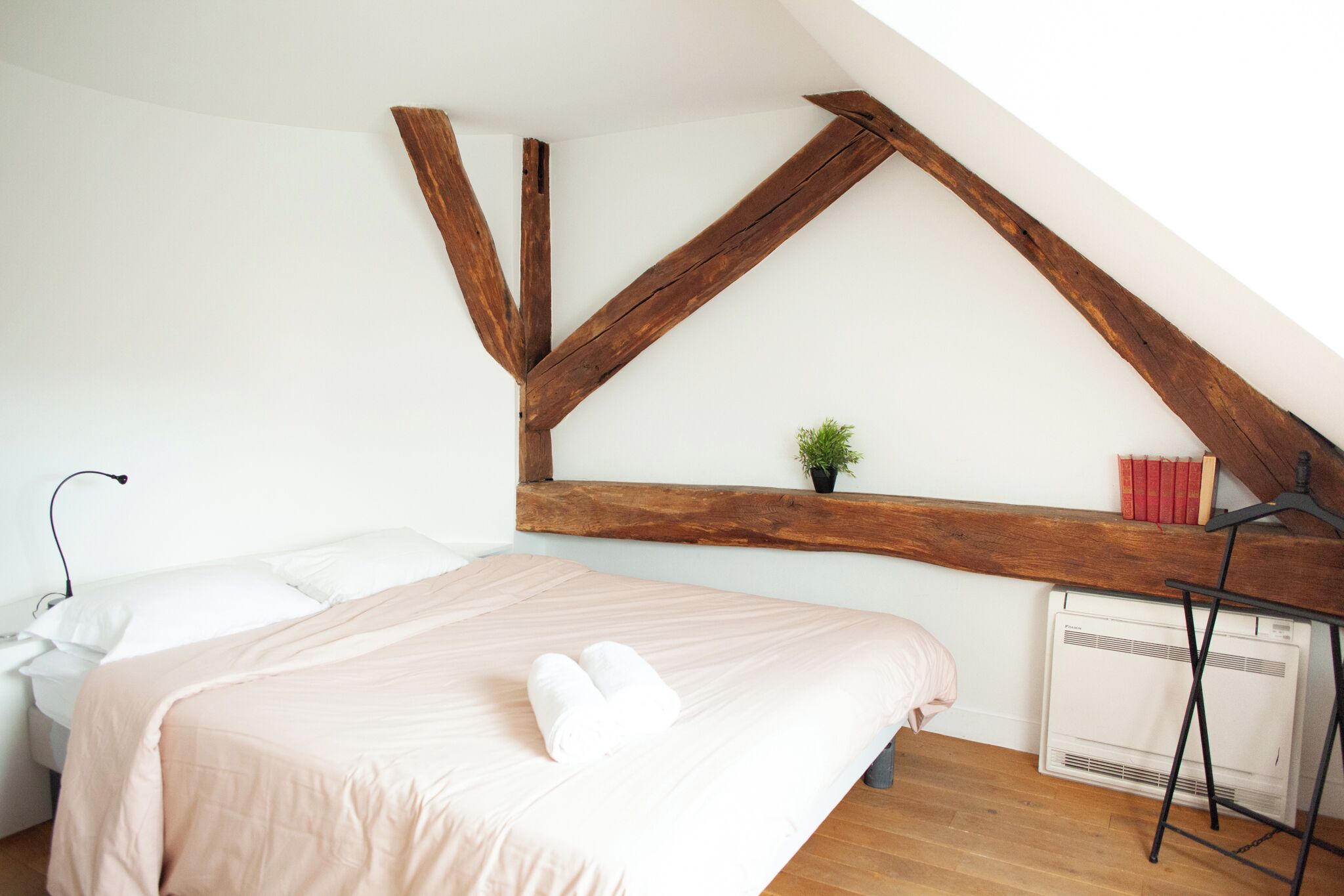 Ein Schlafzimmer mit schrägem Dach, einem Bett mit rosa Bettdecke, einem Nachttisch mit Lampe, einer kleinen Pflanze und einer Heizung.