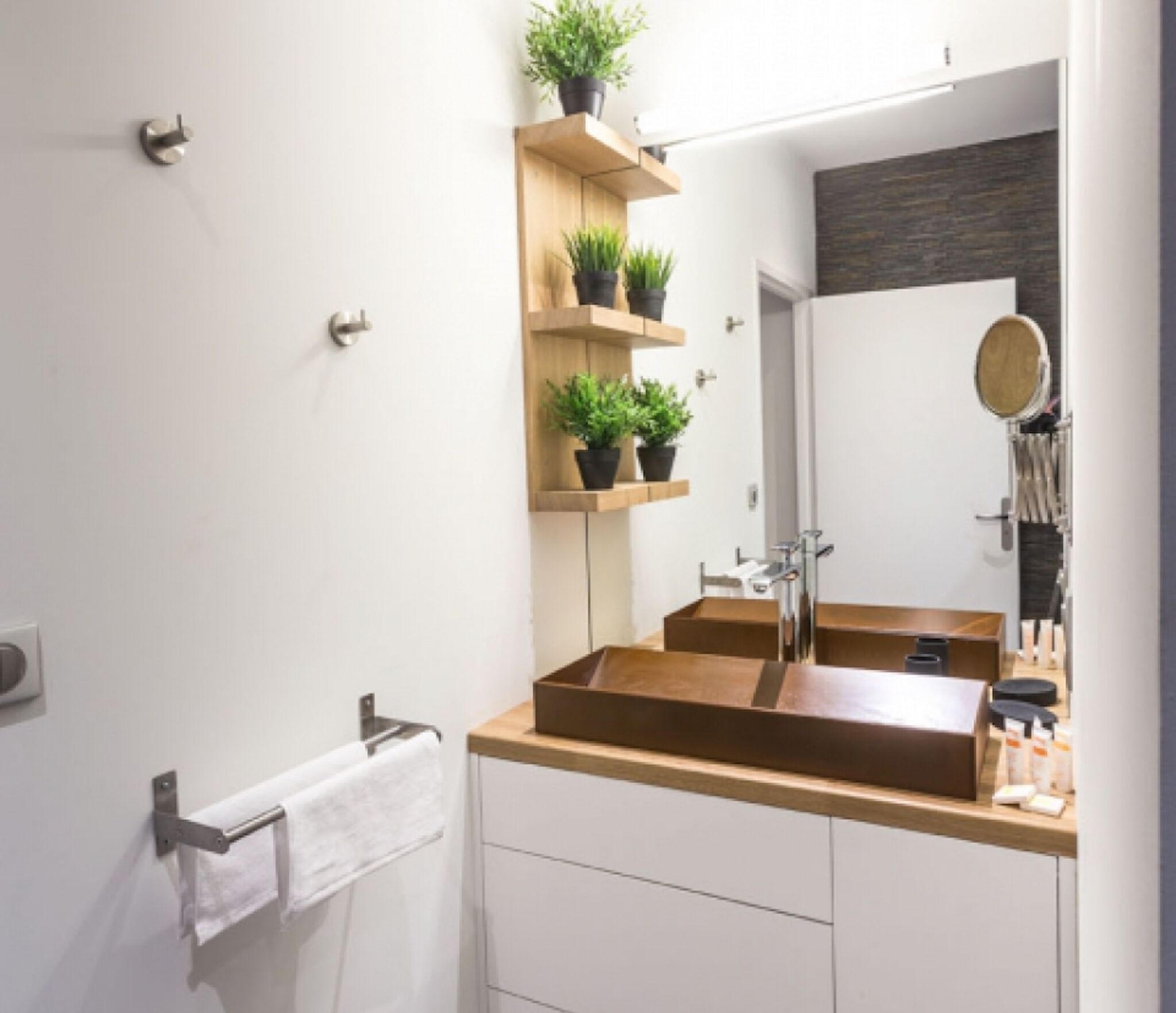 A modern bathroom with a wooden vanity, two sinks, and a large mirror. There are potted plants on a shelf above the mirror.