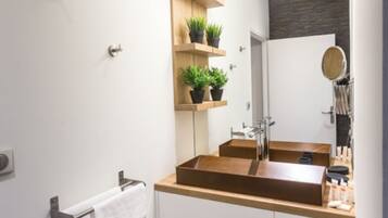 A modern bathroom with a wooden vanity, two sinks, and a large mirror. There are potted plants on a shelf above the mirror.