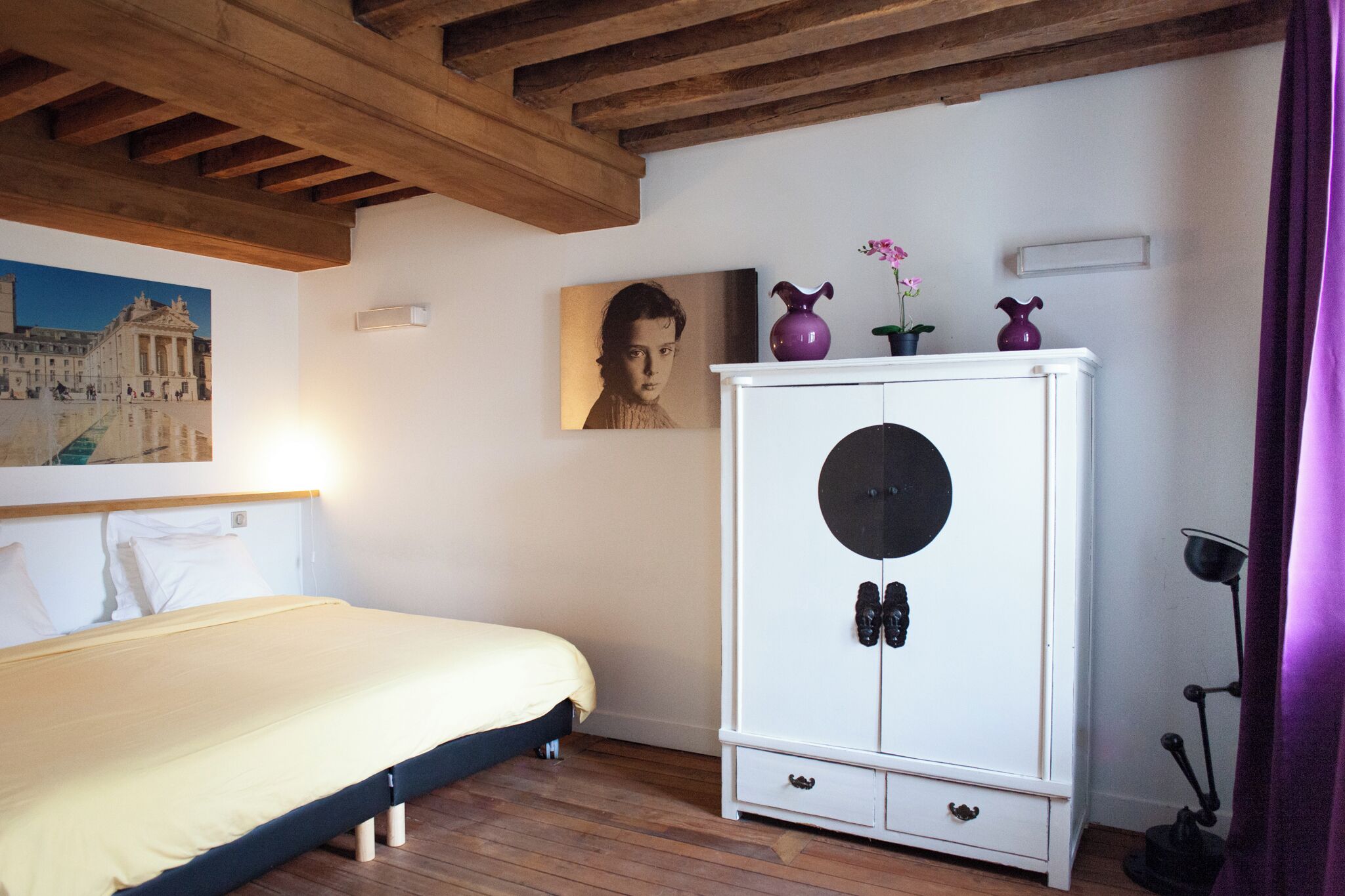 A bedroom with a bed, wooden ceiling beams, a white cabinet with decorative items, and a painting on the wall.