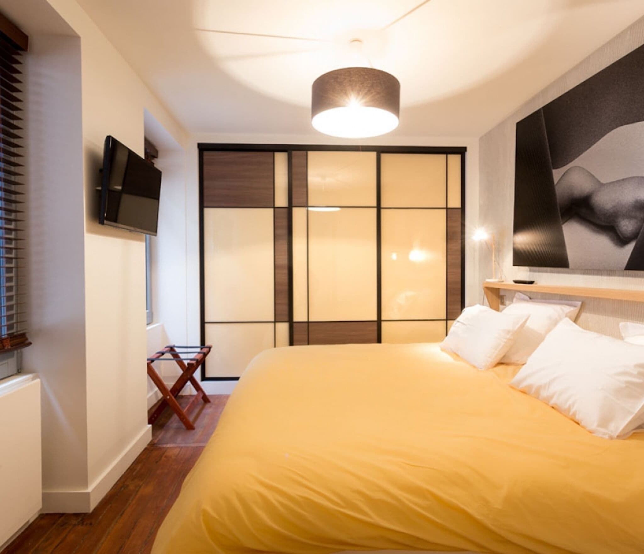 A bedroom with a large bed, a flat-screen TV mounted on the wall, a sliding door with a view, and a ceiling light fixture.