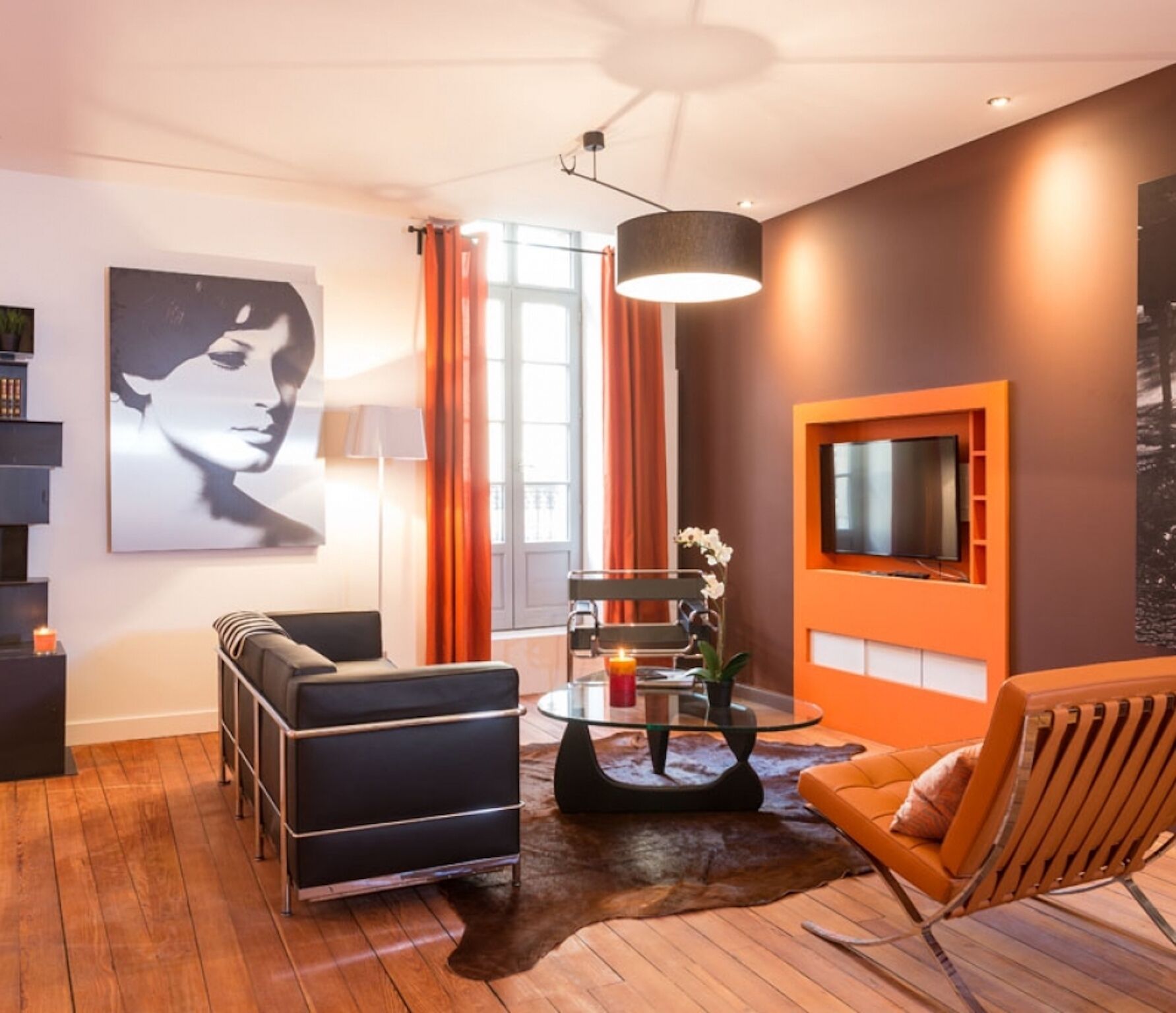 Een moderne woonkamer met een bank, fauteuils, een glazen salontafel en een televisie die aan een oranje muur bevestigd is.