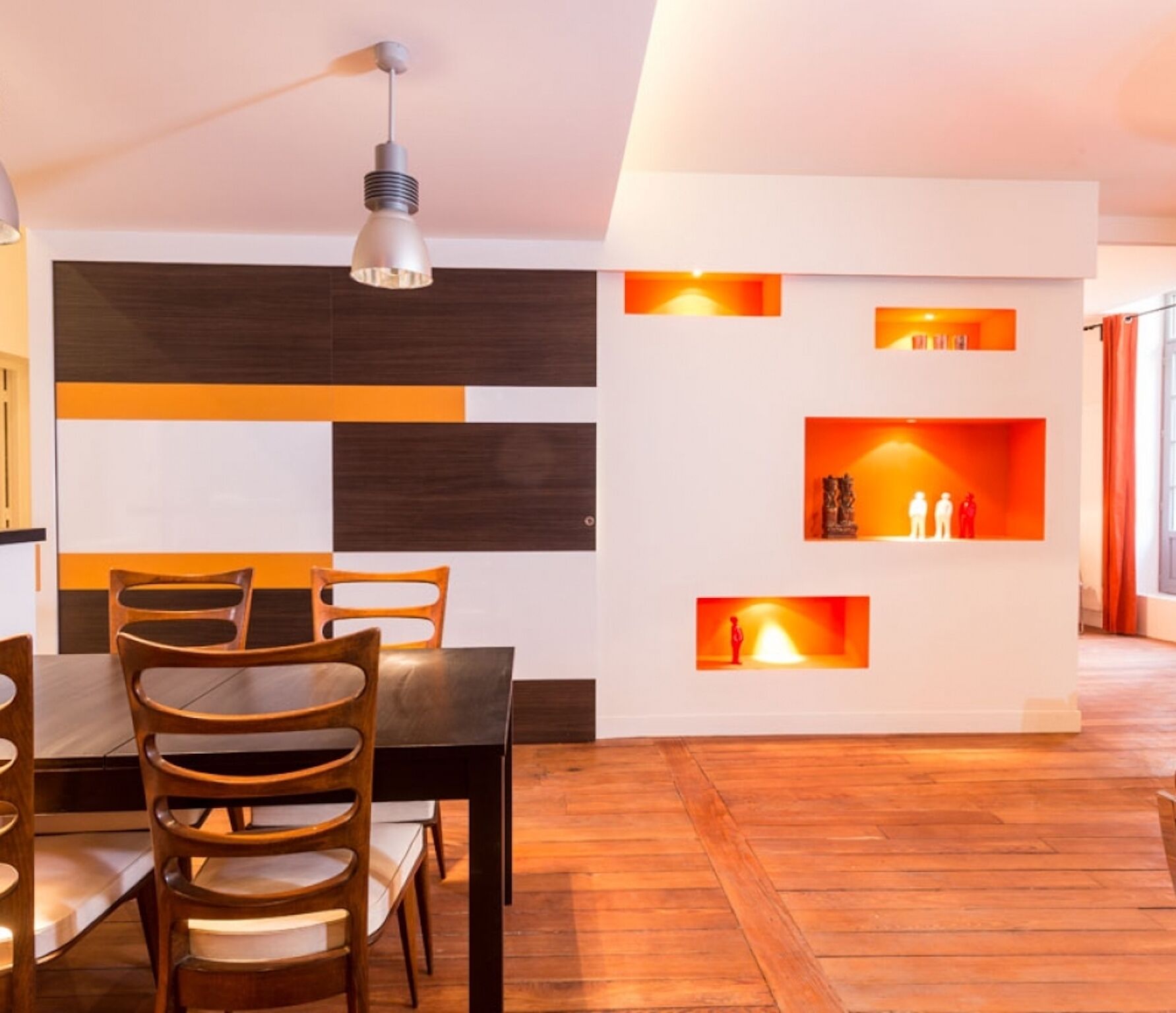 A modern dining area with a wooden table and chairs, a pendant light, and a wall with built-in shelves displaying decorative items.