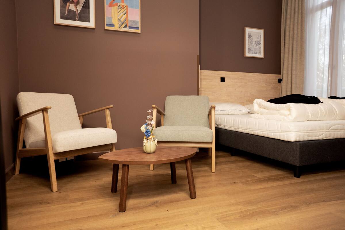 A room with a bed, two chairs, a round wooden table, and a vase with flowers.