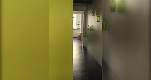 Ein Flur mit modernem Design, der durch einen dunklen Boden und helle Wände gekennzeichnet ist. Es sind grüne Wandleuchten angebracht und im Hintergrund ist eine Küche erkennbar.