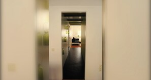 Un couloir au sol noir mène à une pièce où se trouvent un canapé et une lampe.