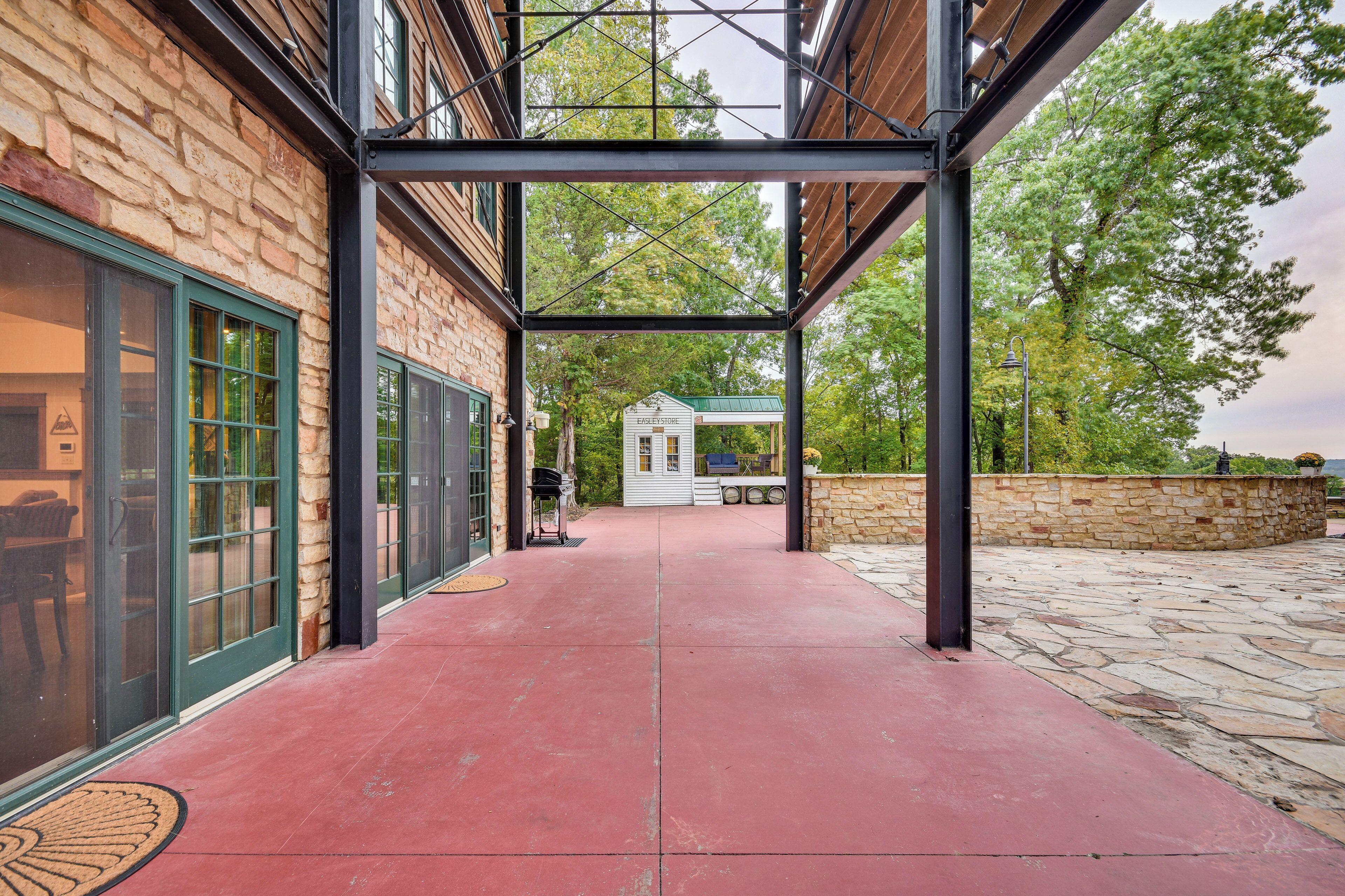 A covered outdoor area with a stone floor, a brick building with large windows, and a small white structure in the background.