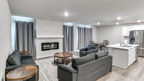 Una sala de estar moderna con un sofá gris, una mesa de centro redonda de madera y una zona de cocina con una isla blanca y electrodomésticos de acero inoxidable.