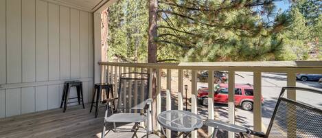 A wooden deck with outdoor furniture, including chairs and a table, overlooking a street with parked cars and trees.