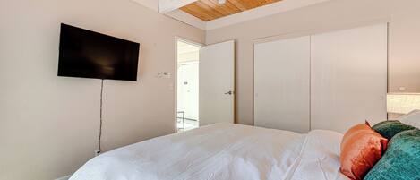 Un dormitorio con techo de madera, un televisor de pantalla plana montado en la pared, una cama con ropa blanca y una puerta que da a un área exterior.