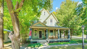 A two-story house with a porch, surrounded by trees and a flag. - Near Lake: Historic Boyne City Home w/ Fire Pit! (Boyne City)