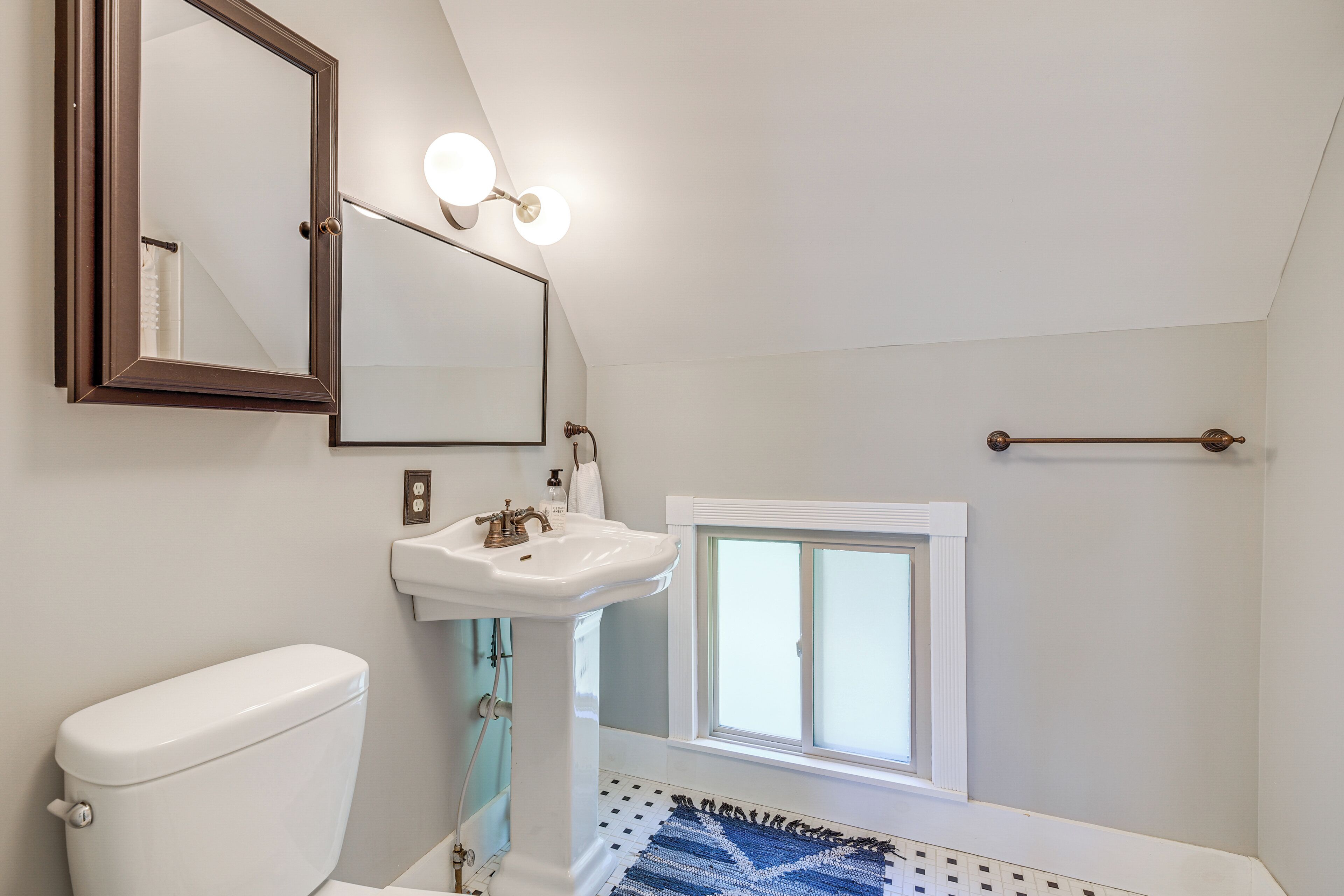 Une salle de bain avec des toilettes blanches, un lavabo sur colonne, un miroir et une fenêtre.
