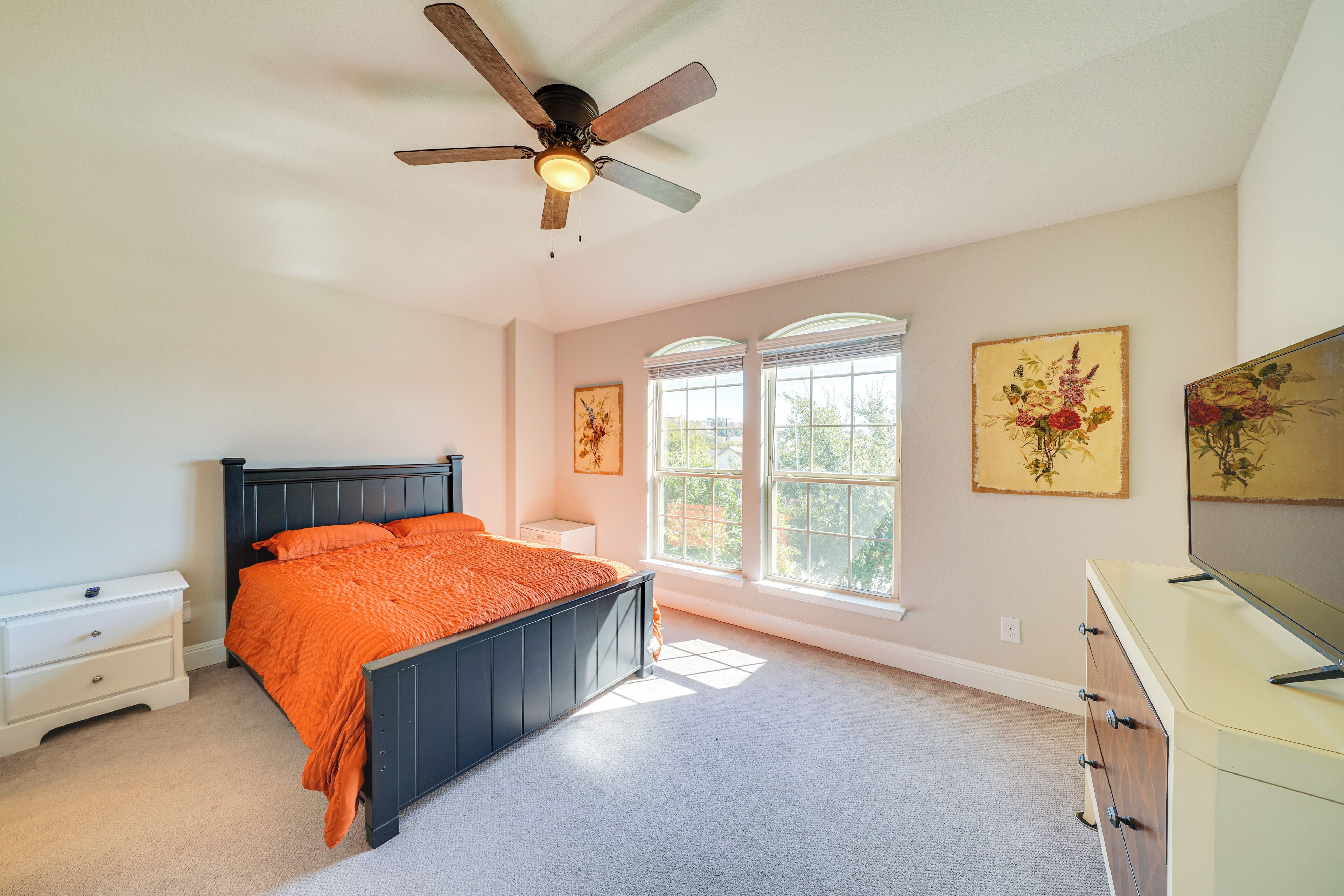 Une chambre à coucher comprenant un lit, une table de chevet, une commode, un ventilateur de plafond et deux fenêtres, les murs étant décorés d’œuvres d’art.