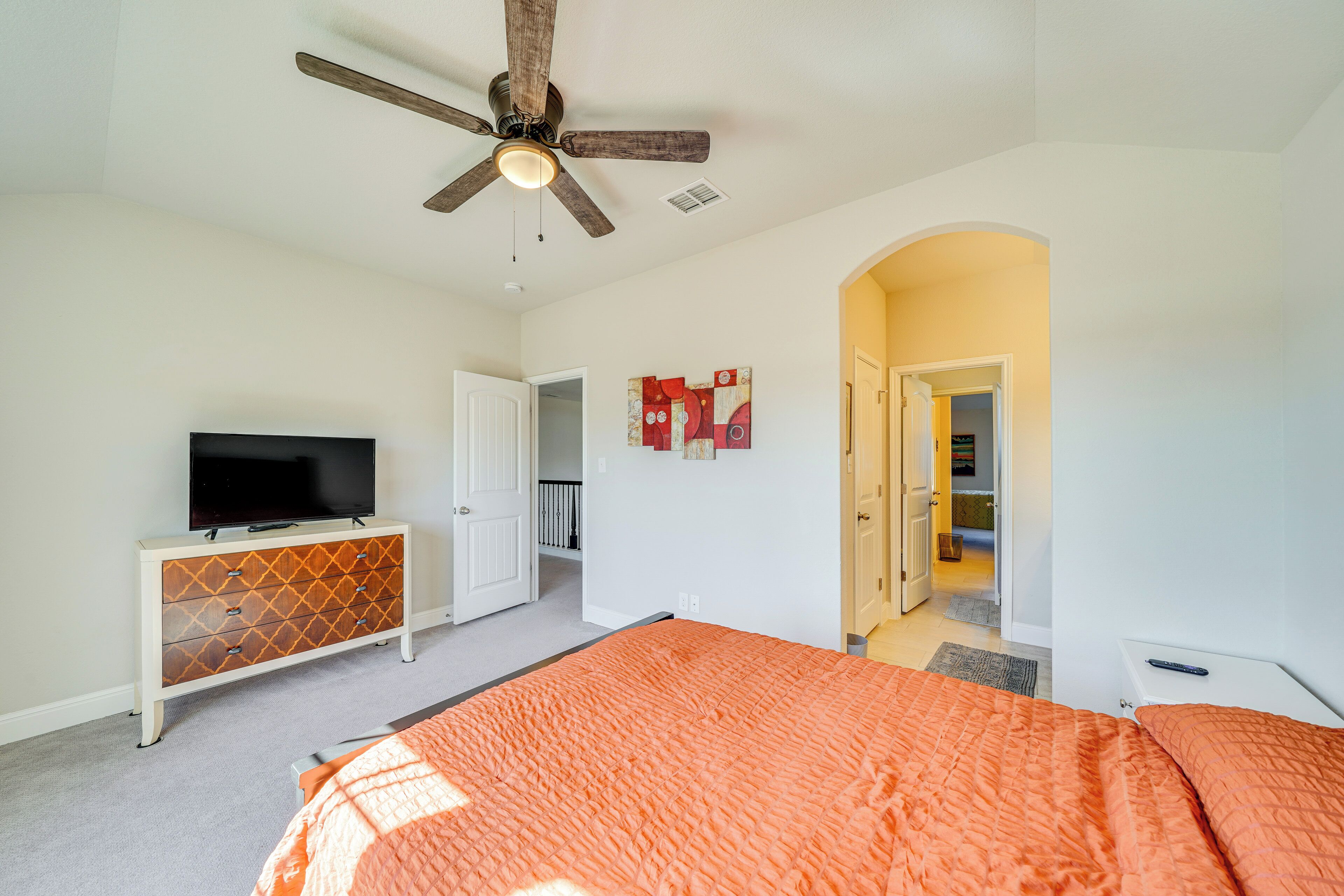 A bedroom with a bed, a TV on a dresser, a ceiling fan, and an open door leading to another room.