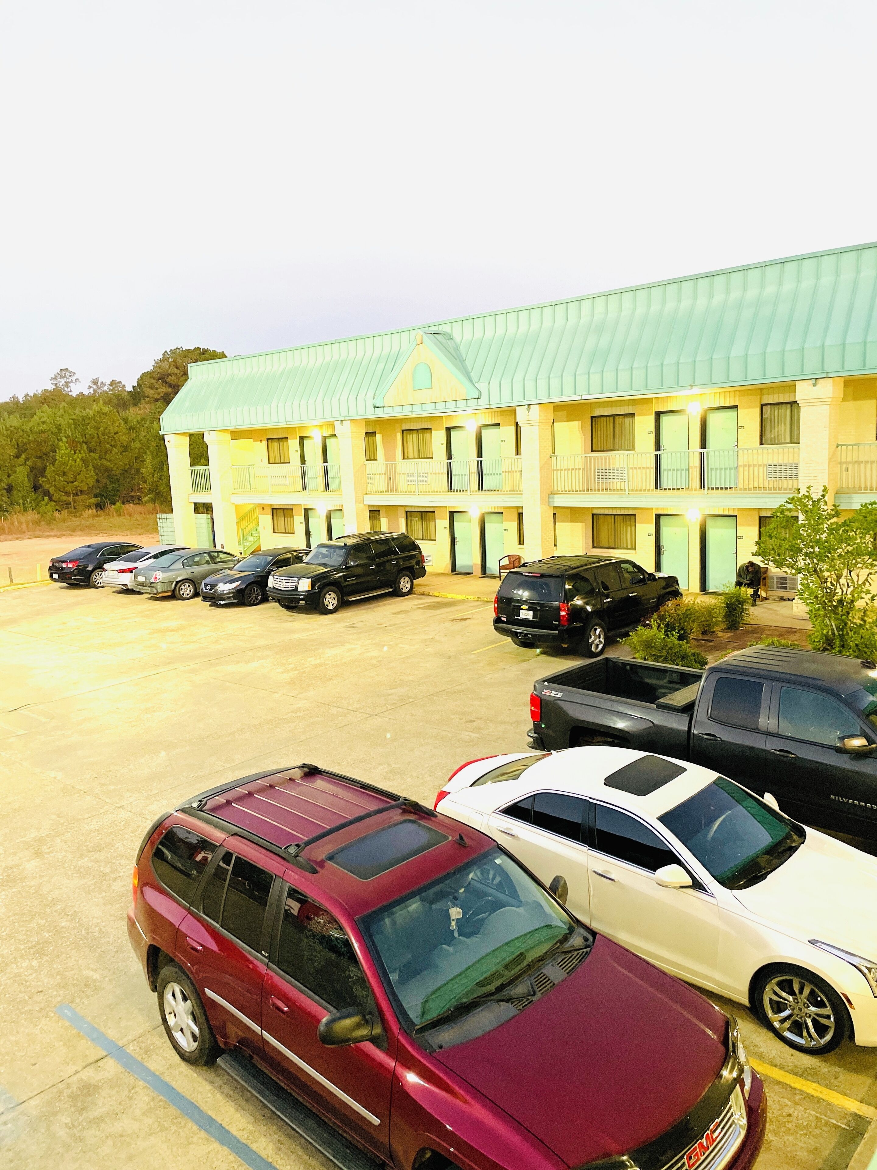 Un motel con techo verde y varios balcones. Se ven varios coches aparcados en la entrada, incluyendo un SUV color burdeos y un sedán blanco.