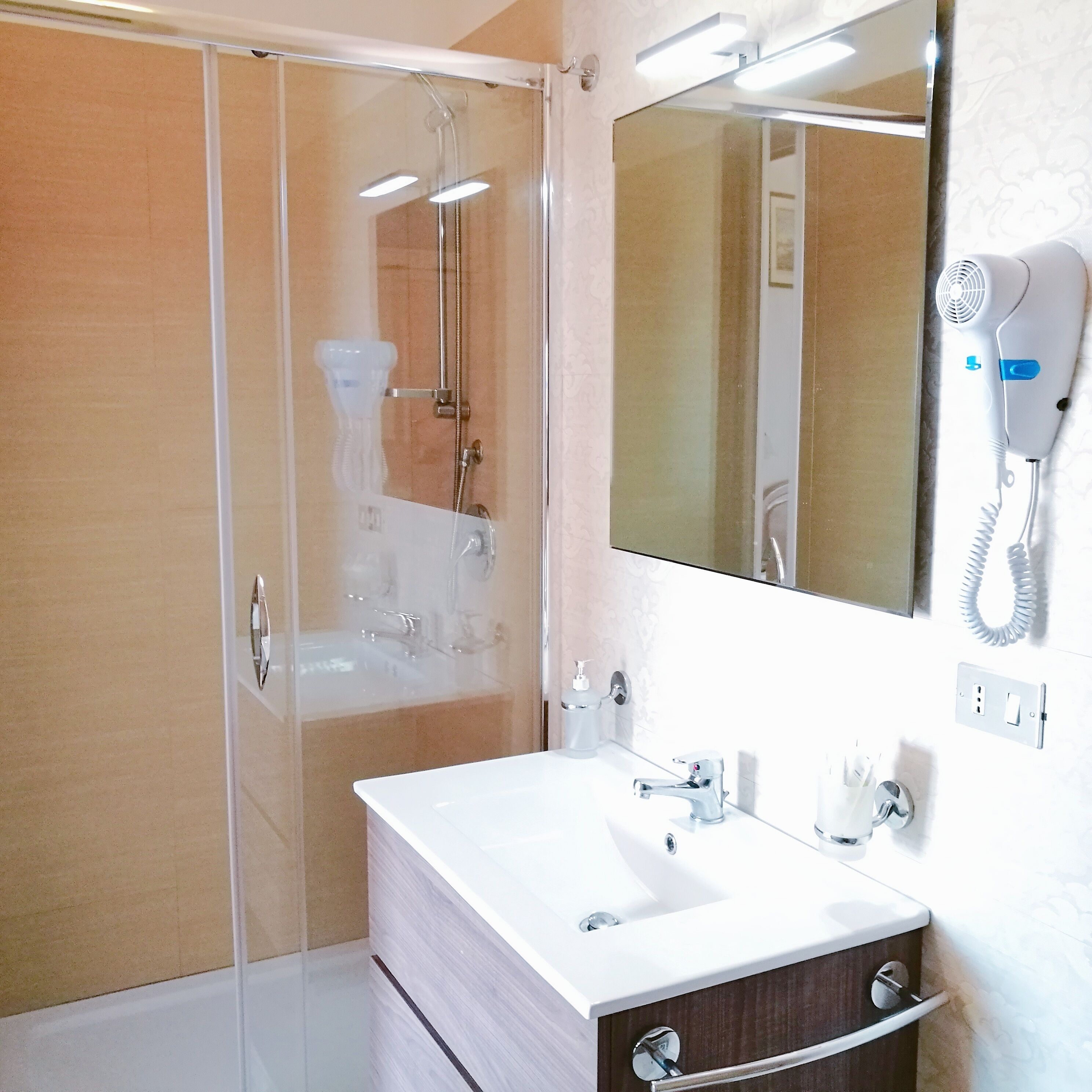 A modern bathroom with a glass shower enclosure, a white sink with a mirror above it, and a hairdryer mounted on the wall.