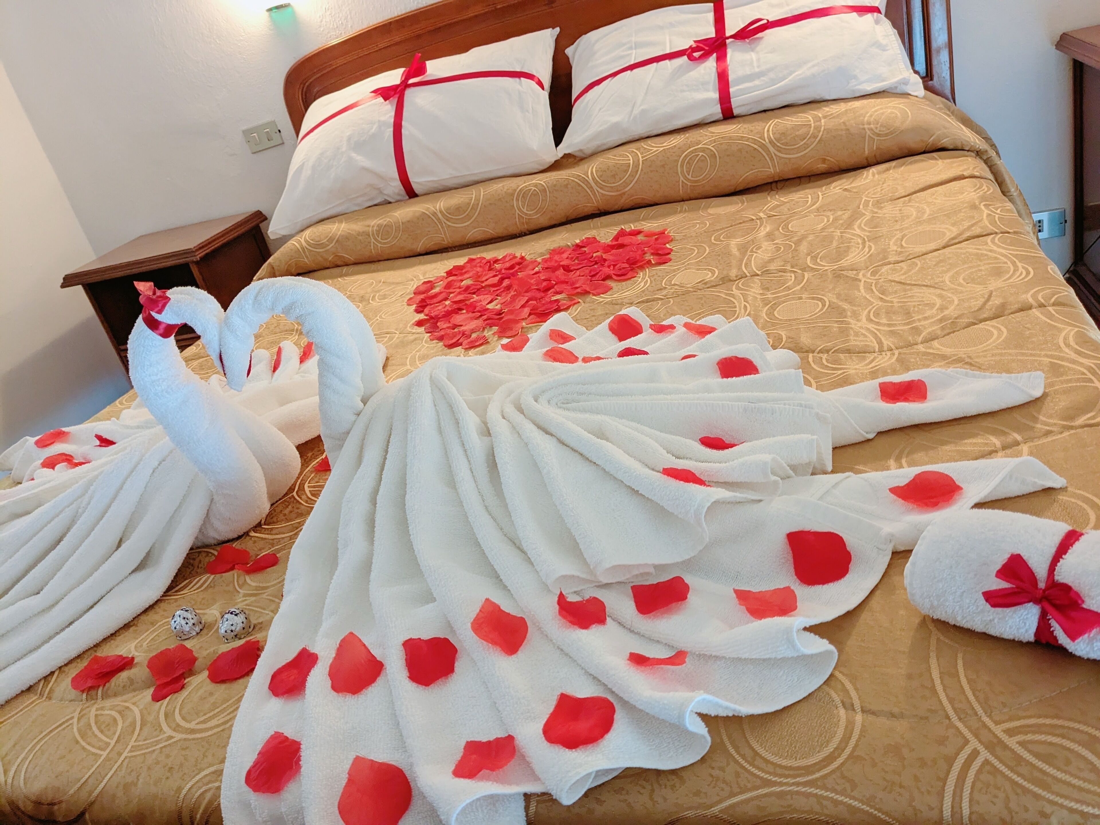 A bed with a heart-shaped decoration and rose petals, a wooden headboard, and white pillows with red ribbons.