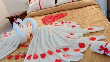 A bed with a heart-shaped decoration and rose petals, a wooden headboard, and white pillows with red ribbons.