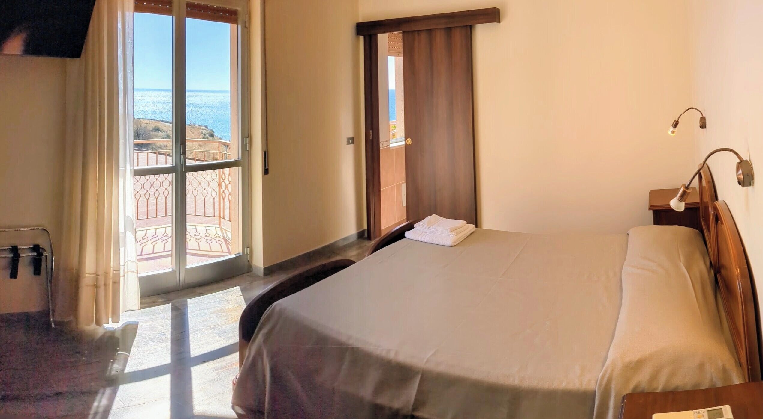 A hotel room with a bed, a nightstand, a lamp, and a view of the sea through a glass door.