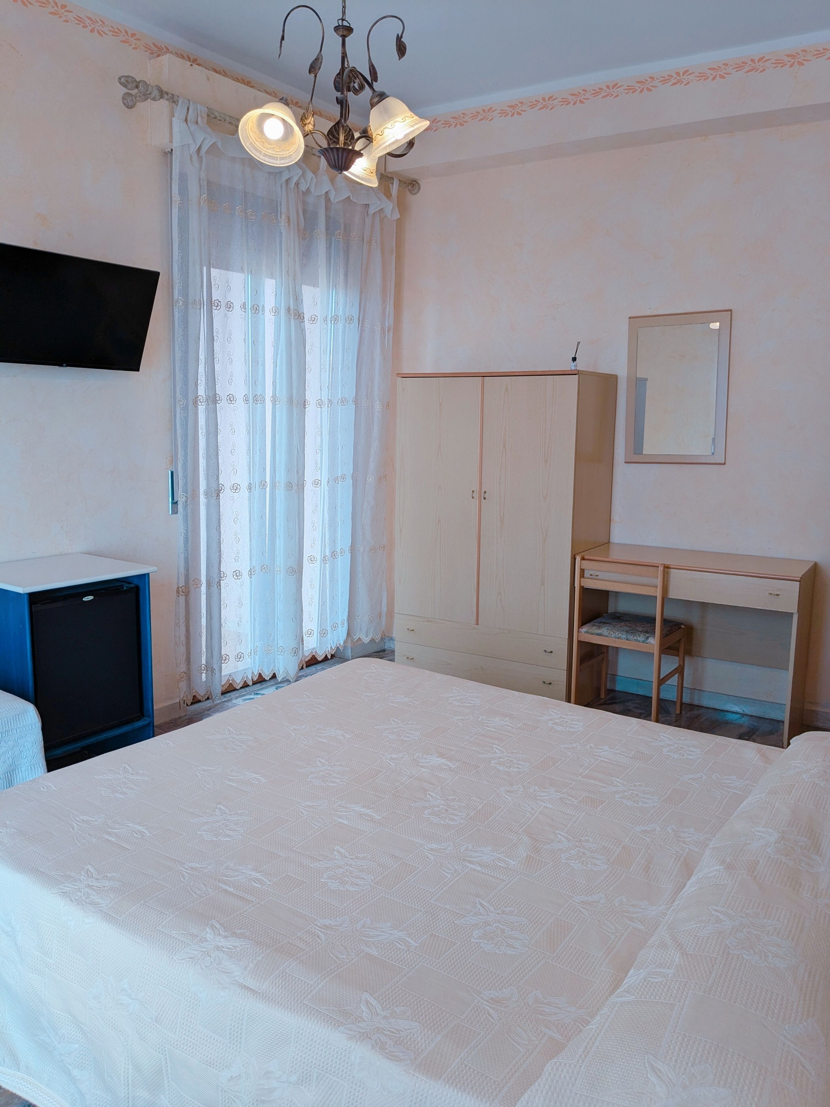 Un dormitorio con cama, armario, escritorio, silla, televisor y ventana con cortinas.