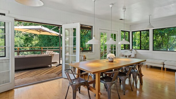 A spacious dining area with a wooden table and metal chairs, large windows with a view of greenery, and pendant lighting.