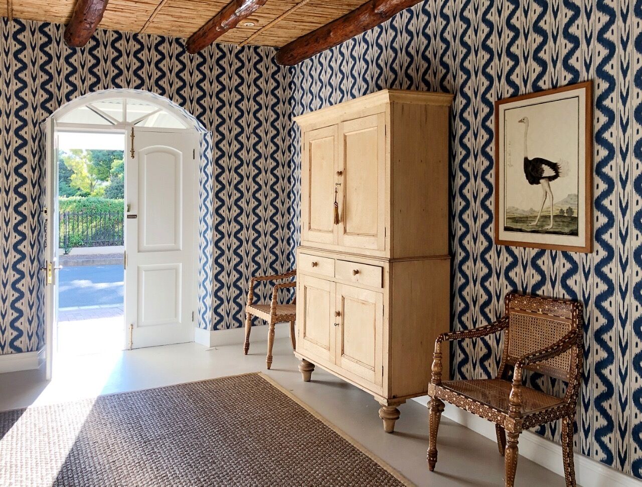 A room with patterned wallpaper, a wooden cabinet, two wicker chairs, and a framed picture on the wall.