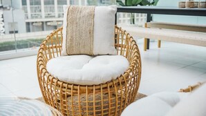 Een rieten stoel met een witte kussen en een gebreid plaid, geplaatst op een geweven mat.