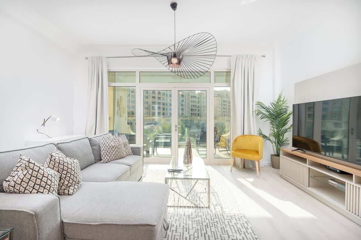 Un salon moderne avec un canapé gris, un fauteuil jaune, une table basse en verre et un téléviseur posé sur un meuble en bois.