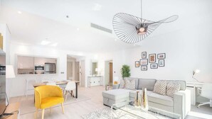 Ein modernes Wohnzimmer mit einer Couch, einem Glastisch und einem gelben Sessel. Im Hintergrund ist eine Küche mit Kühlschrank und Mikrowelle zu sehen.