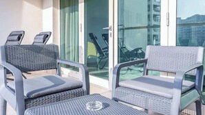 Ein Balkon mit zwei Korbstühlen und einem kleinen Tisch, eine Glastür, die sich öffnen lässt, sowie ein Blick auf ein Gebäude und Berge in der Ferne.