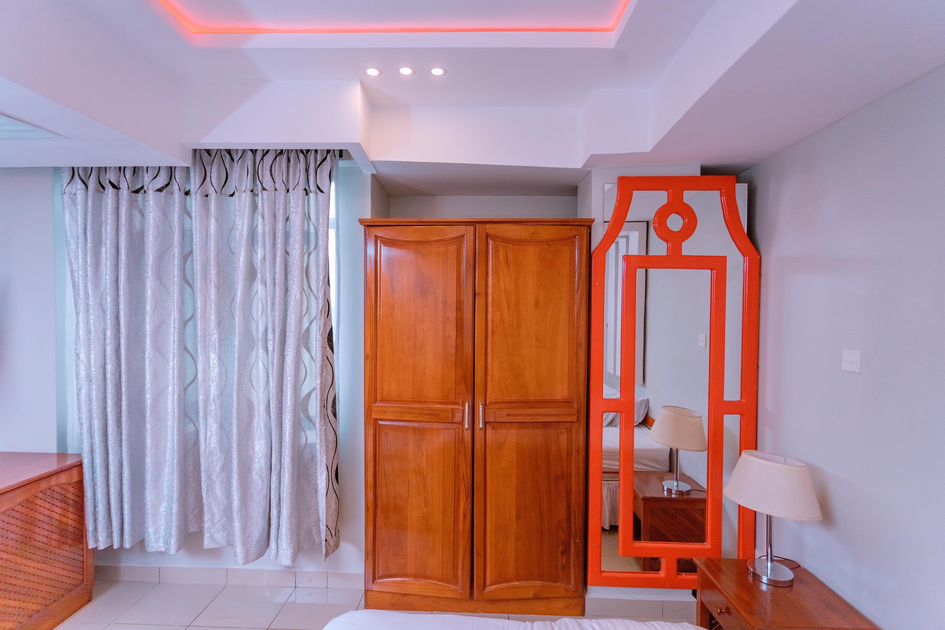 A bedroom with a wardrobe, a mirror, a bedside table, and a lamp.