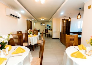 Un comedor de hotel con mesas dispuestas para los huéspedes, una recepción y una pantalla de televisión al fondo.