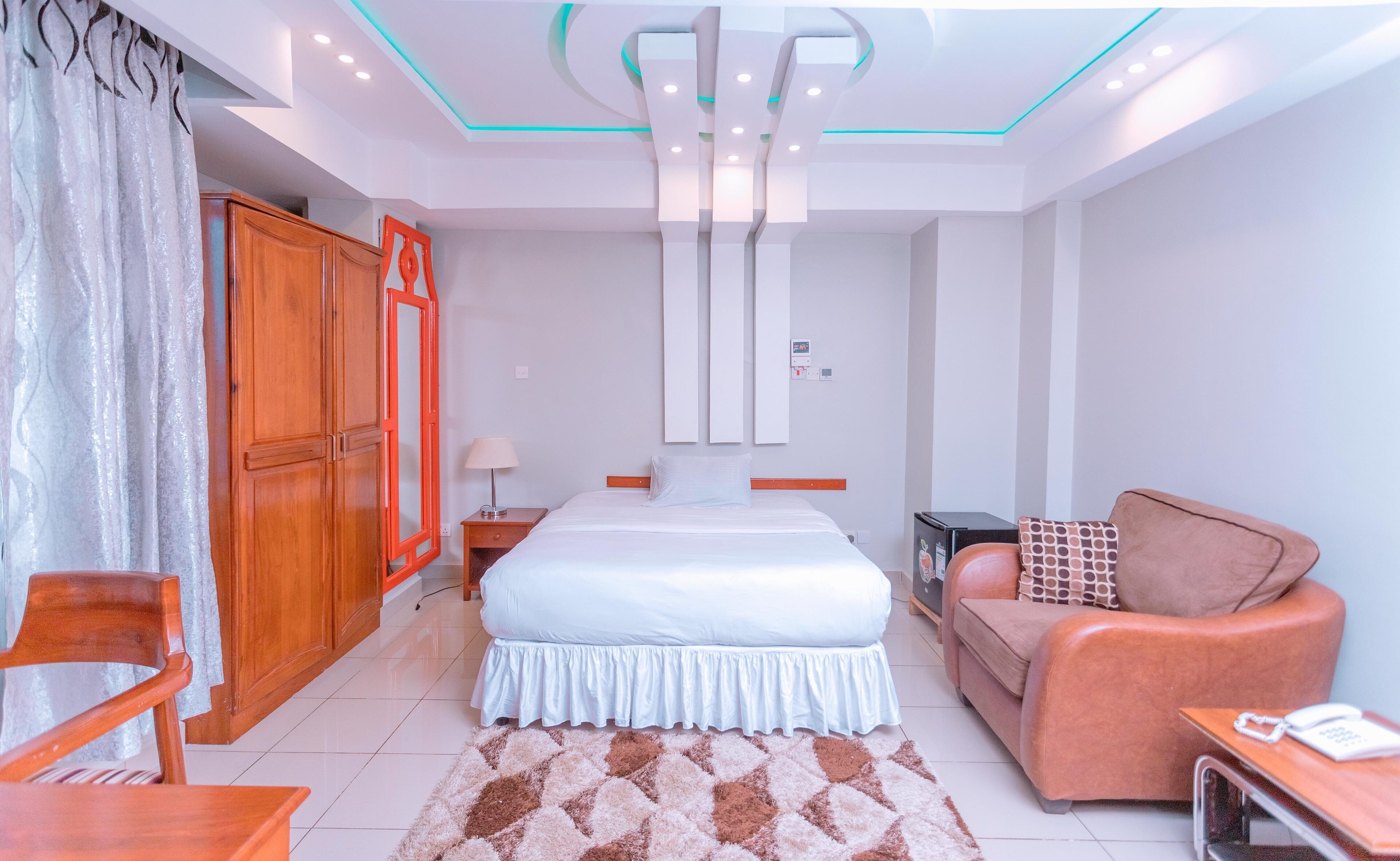 A hotel room with a bed, a sofa, a wooden wardrobe, a desk with a phone, and a patterned rug.