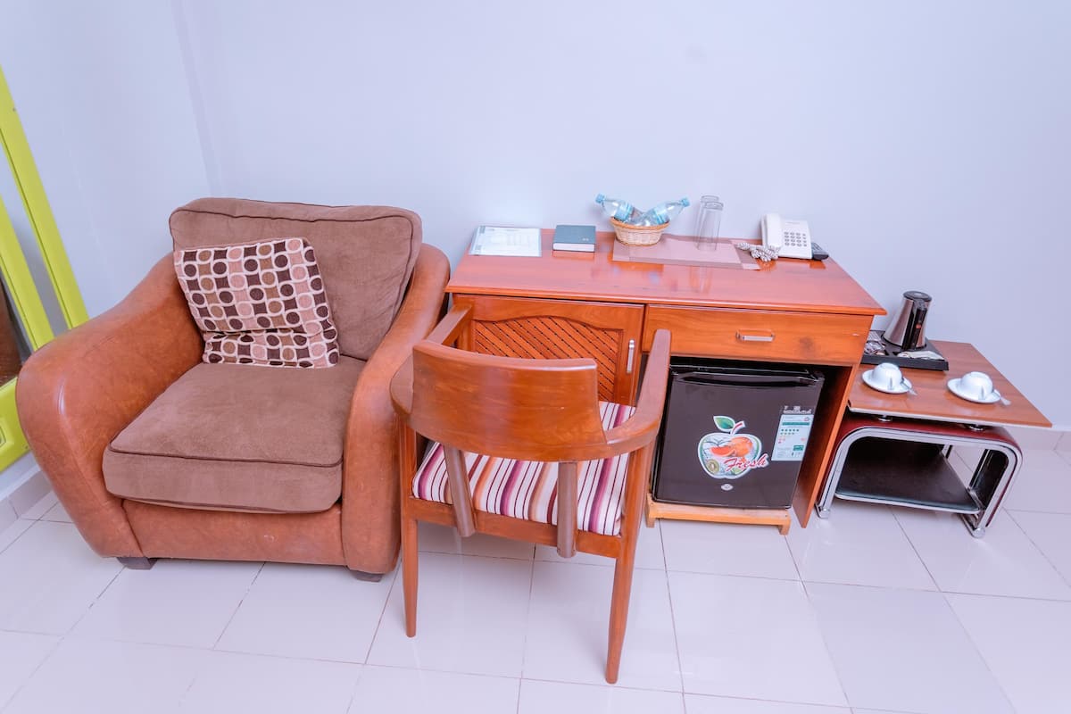 A compact living space with a brown sofa, a wooden chair, a small desk with a phone, a basket of bottled water, and a mini refrigerator.