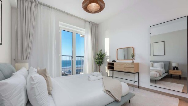 A modern bedroom with a large bed, a bedside table, a mirror, and a view of the outdoors. - StayShort - Port De La Mer 1BR for 2, steps from the beach (Dubai)
