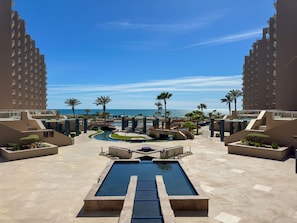 A resort pool area with a central pool, palm trees, and a view of the ocean. - Oceanfront Vibes Iconic Beachside Getaway (Puerto Peñasco)