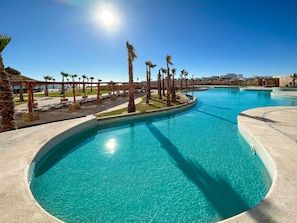 A large outdoor swimming pool with clear blue water, surrounded by palm trees and a covered seating area. - Oceanfront Vibes Iconic Beachside Getaway (Puerto Peñasco)