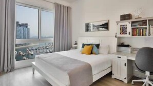 A modern bedroom with a large bed, a desk, and a chair. There is a window with curtains and a view of buildings. - StayShort - Superb 3BR in Palm Jumeirah With Unmatched Views! (Marina/JBR)