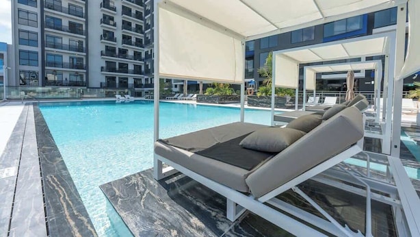 Area relax a bordo piscina con una sdraio, piscina cristallina e un edificio moderno sullo sfondo.