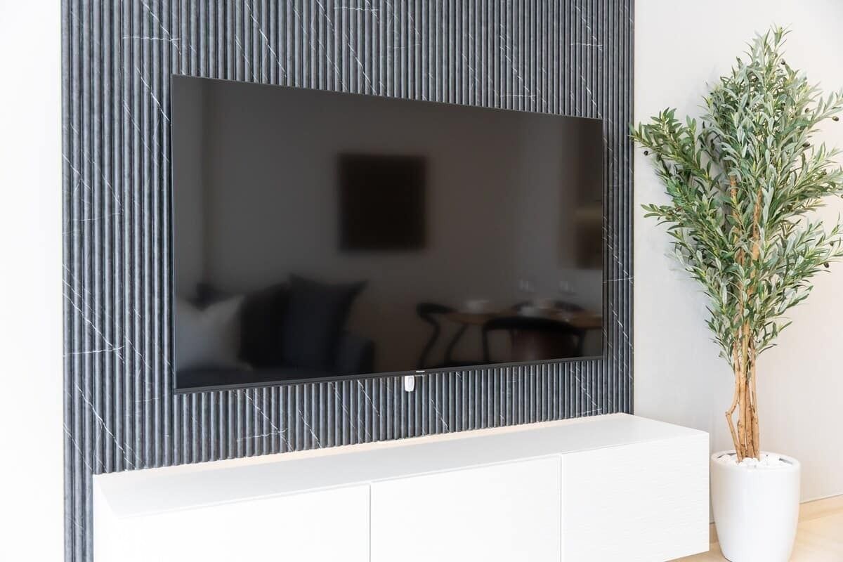 Un salon moderne avec un grand téléviseur à écran plat fixé à un mur texturé, une console blanche en dessous et une plante en pot sur la droite.